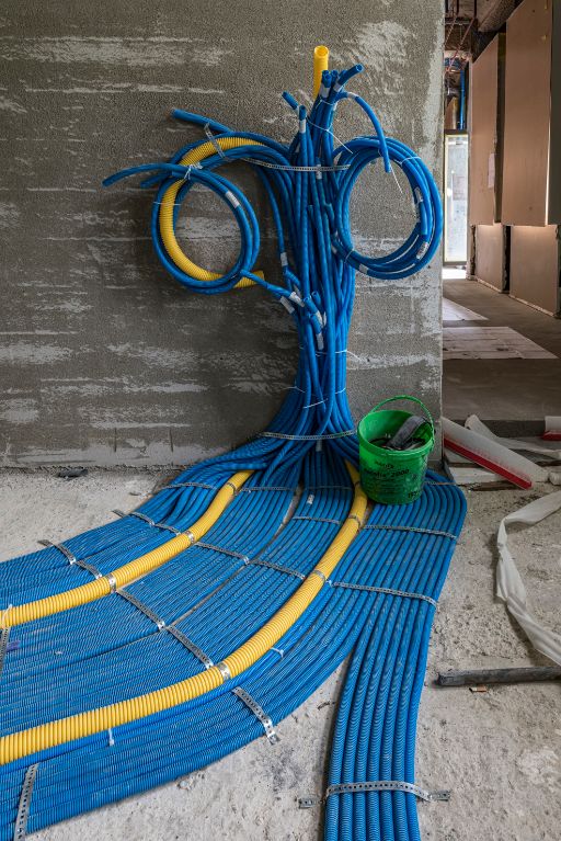 Bündel aus blauen und gelben Kabelrohren, die an einer Wand hochlaufen und sich wie Äste eines Baumes verzweigen. Auf dem Boden verlaufen die Rohre geordnet in Richtung Raum.